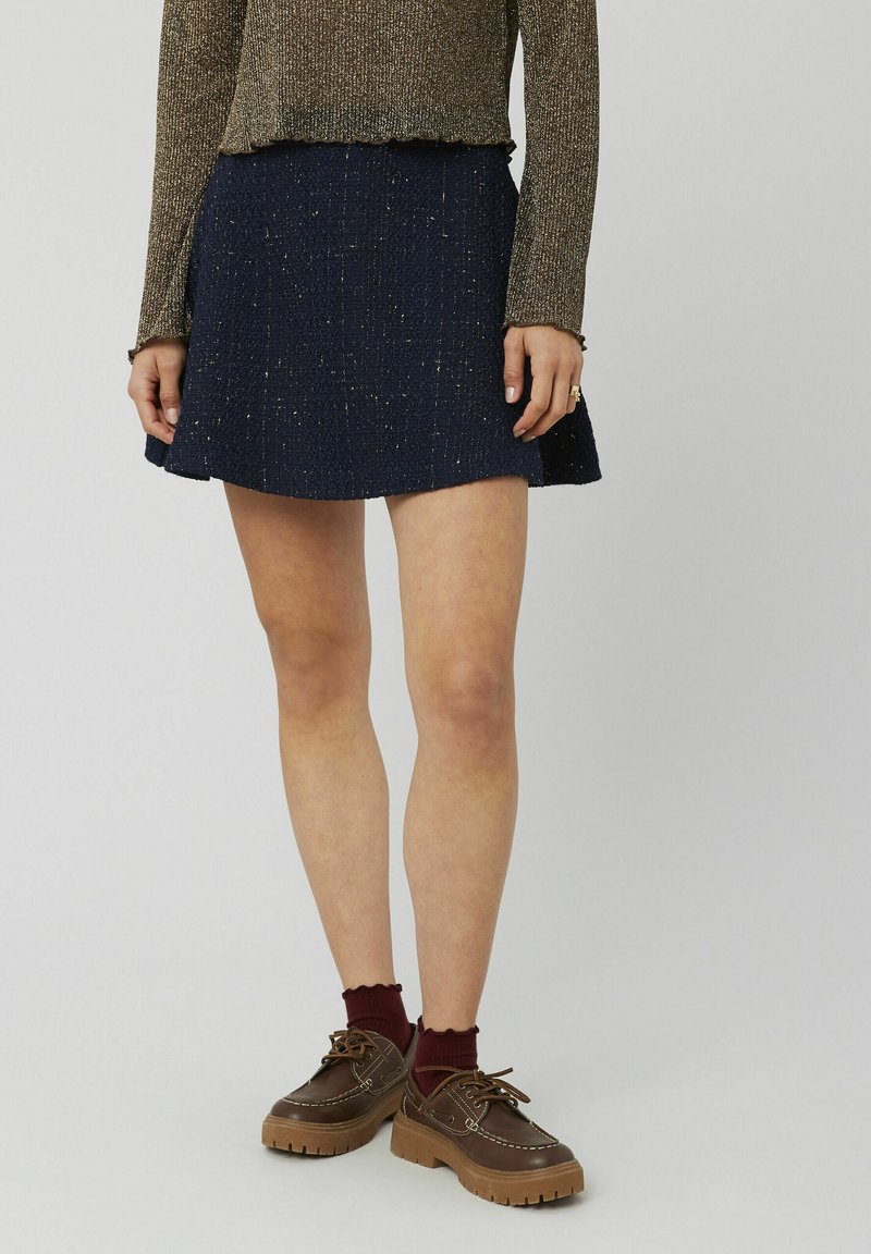 Jupe bleu marine en tissu texturé avec un ourlet évasé. Associée à des chaussures marron et des chaussettes bordeaux, ajoutant du contraste et des détails.