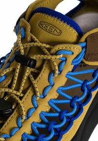 Zapato de gamuza amarilla con cordones elásticos azules, hardware negro y suela texturizada. Presenta un collar acolchado y la marca Keen en el lateral.