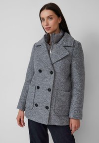 Femme portant un manteau en laine grise texturé avec des boutons noirs sur une veste matelassée zippée et un pantalon foncé, se tenant contre un fond uni.