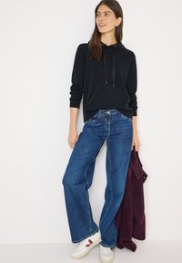 Czarna bluza z kapturem z przednią kieszenią, zestawiona z luźnymi niebieskimi dżinsami. Białe trampki z czerwonymi akcentami. Śliwkowy sweter zarzucony na ramię.