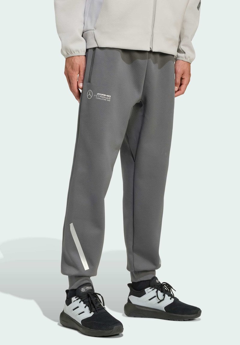 Graue Sweatpants aus weichem Material, mit einem weißen Logo, einem reflektierenden Seitenstreifen und einem elastischen Bund. Kombiniert mit schwarzen Turnschuhen.