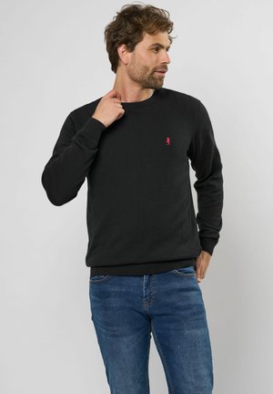 Zwart gebreid sweatshirt met een ronde hals en geribbelde cuffs, met een klein rood logo op de borst. Gemaakt in combinatie met blauwe denim jeans.