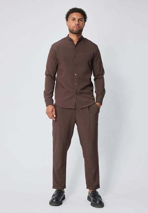 Chemise marron à manches longues avec col mandarin, associée à un pantalon assorti. Le tissu semble lisse et le style est ajusté. Des chaussures noires complètent le look.