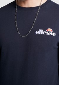 Mörkblå sweatshirt med ribbad rund halsringning, med vit broderad "ellesse"-logotyp och en flerfärgad detalj. Silverkedja syns.
