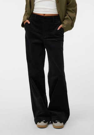 Personne portant un pantalon large en velours côtelé noir, des baskets blanches, un top court blanc et une veste verte boutonnée, les mains dans les poches.