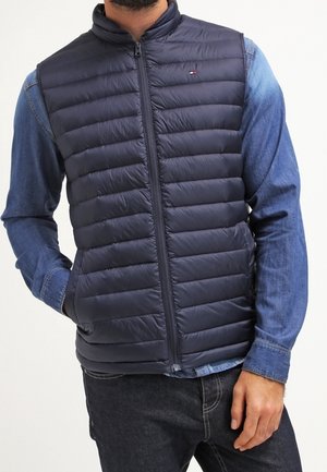Man wearing navy blue quilted sleeveless vest over denim shirt with hands partially in pockets, standing against white background.