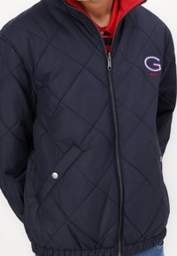 Mann trägt eine marineblaue gesteppte Zip-Jacke mit rotem Innenfutter und "G"-Logo auf der Brust.