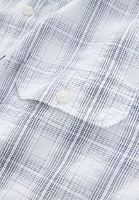 Camicia a maniche corte a quadri in bianco e blu navy; presenta un tessuto a trama, chiusura con bottoni e una tasca sul petto con patta.