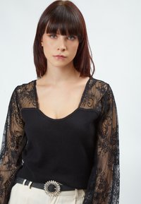 Schwarzes Oberteil mit Rundhalsausschnitt und langen Spitzenärmeln, das mit floralen Mustern verziert ist. Kombiniert mit einem schwarzen Gürtel mit einer dekorativen Strassschnalle.