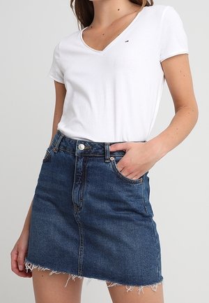 Mujer con camiseta blanca de manga corta y escote en V y falda vaquera azul de cintura alta con borde deshilachado, mano en el bolsillo de la falda.
