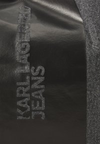 Svart denimstoff med teksturert bokstavering "KARL LAGERFELD JEANS" i preget detalj, utfylt med en kontrasterende grå stoffkant.
