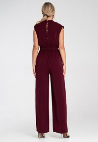 Burgundowy kombinezon z wysokim dekoltem, krótkimi rękawami, elastycznym pasem, szerokimi nogawkami i zapięciem na guziki z tyłu. Gładka tekstura materiału.