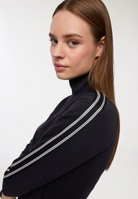 Giovane donna con pelle chiara e capelli castani lisci, che indossa un dolcevita nero con strisce bianche sulle maniche, mentre guarda leggermente di lato.