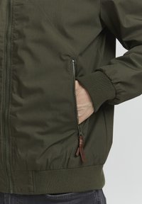 Maslinastozeleni bomber jakna s glatkom tkaninom, rebrastim manžetama i bočnim džepom sa zatvaračem i kožnom uzicom.