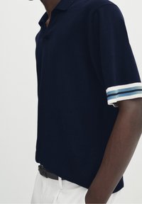 Homme portant un polo à manches courtes bleu marine avec des poignets rayés blancs et bleus, et un pantalon blanc avec une ceinture noire, sur fond uni.