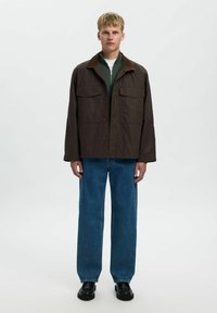 Giacca da campo marrone con colletto marrone, due tasche sul petto, indossata sopra una maglietta verde, abbinata a jeans blu larghi e scarpe nere.