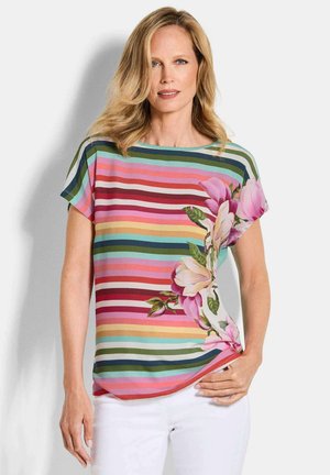 Femme portant un chemisier rayé coloré avec un grand imprimé floral rose sur un côté, associée à un pantalon blanc, se tenant contre un fond uni.