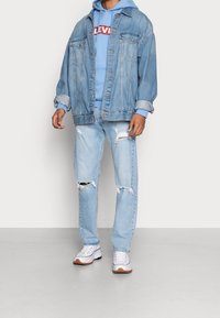 Ljusblå denimjacka över en ljusblå huvtröja med logotyp, matchad med slitna ljusblå jeans och vita sneakers med bruna detaljer.