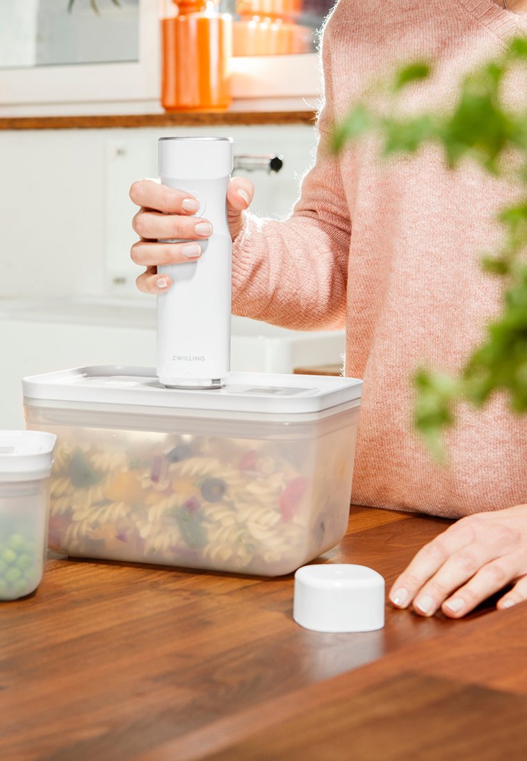 Weißer Lebensmittel-Vakuumpumpe in der Hand, versiegelt einen transparenten Behälter mit mehrfarbiger Pasta. Holzoberfläche des Tisches, unscharfer grünlicher Hintergrund.