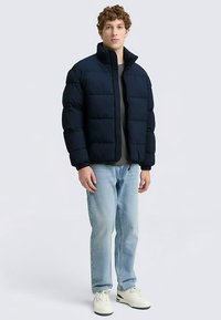 Temno modra puhovka z visokim ovratnikom, z vzorcem šivov, sprednja zadrga in rahlo ohlapen kroj; nošena čez sivo majico in kavbojke.