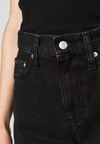 Jeans de mezclilla negra con una textura suave, que presentan herrajes plateados y un botón con el logo de CK. Incluyen cinco bolsillos y un diseño de trabillas para cinturón.