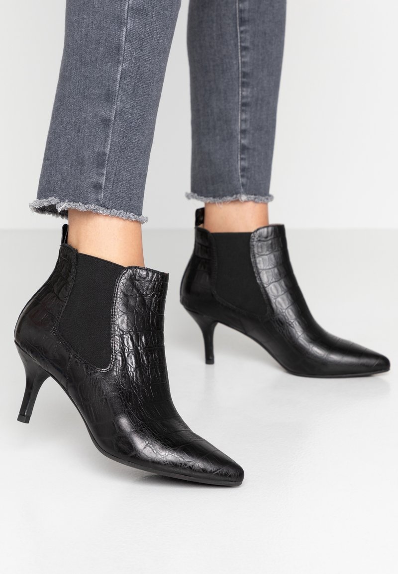 Bottes bottines en cuir noir à motif crocodile avec bouts pointus et talons kitten, portées avec un jean skinny gris à ourlets effilochés.