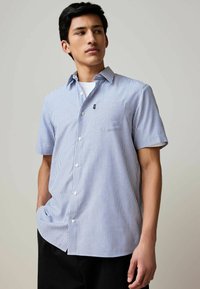 Chemise à manches courtes à boutons, rayée bleu et blanc, avec une poche poitrine et un col pointu. Le tissu semble léger.
