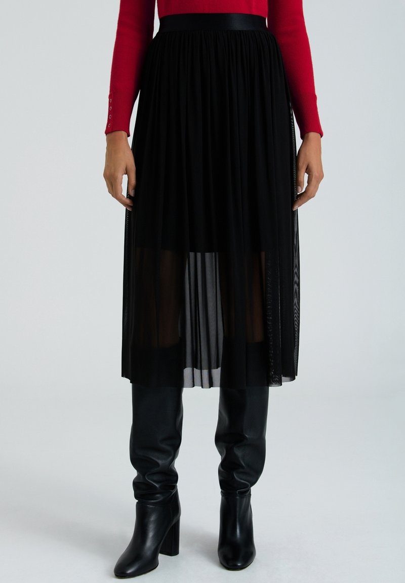 Jupe plissée noire transparente de longueur moyenne portée sur des bottes en cuir noires montantes jusqu’aux genoux, portée avec un haut rouge à manches longues.