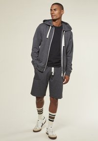 Man wearing dark gray zip-up hoodie, matching shorts, black t-shirt, striped socks, and white sneakers, standing against plain background.