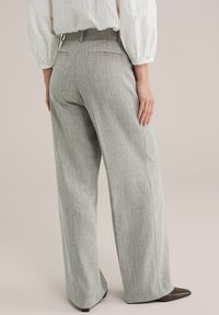 Femeie purtând pantaloni largi cu talie înaltă, model herringbone gri, și o bluză albă brodată, stând cu o mână sprijinită pe șold.