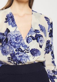 Blusa floreale con scollatura a V profonda, caratterizzata da motivi floreali blu e bianchi su sfondo crema. Tessuto leggero con una consistenza morbida.