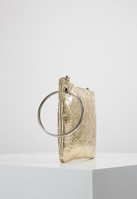 Złota metaliczna torebka o marszczonej fakturze, prostokątnym kształcie i dużym okrągłym metalowym uchwycie, z zamknięciem na zamek błyskawiczny na górze.