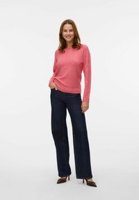 Un pull en laine rose avec un col rond et des manches longues, associé à un jean flare bleu foncé. Des chaussures à talons marron avec des embellissements.