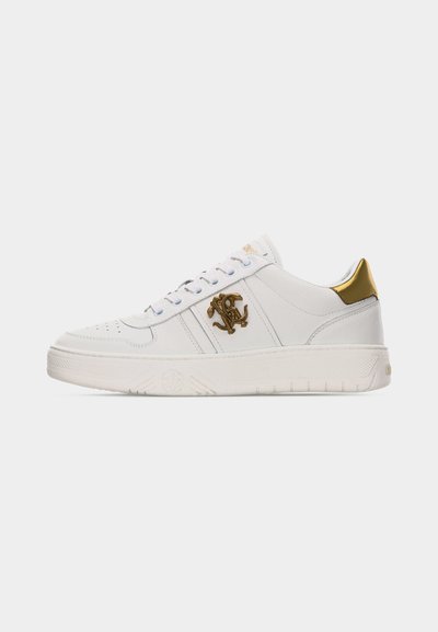 Άσπρο χαμηλό sneaker με χρυσό λογότυπο στο πλάι και χρυσή ταμπέλα στη φτέρνα, άσπρα κορδόνια και υφή στη σόλα, σε απλό φόντο.