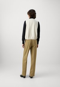 Slonovinová bezrukávová vesta s žebrovaným límcem, vybavená elastickým lemem, v kombinaci s béžovými kalhotami s vysokým pasem a černými botami.