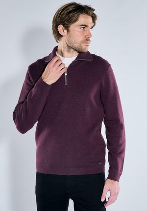 Mann trägt einen burgunderfarbenen Reißverschluss-Pullover über einem weißen Hemd, schaut nach rechts und berührt mit einer Hand den Reißverschluss.