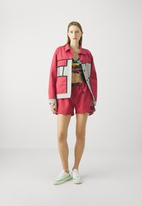 Rød denimjakke med grå detaljer, mønstrete crop topp, røde shorts og hvite joggesko med grønn trim. Uformell antrekk med enkle design.