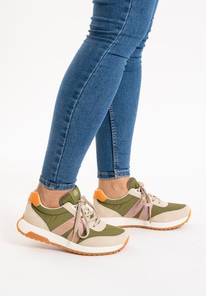 Person wearing slim blue jeans and green, beige, and orange casual sneakers with laces, standing on a white background.