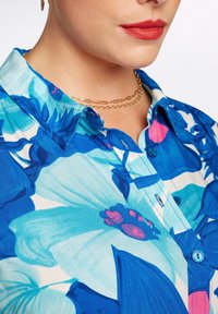 Blå blommig skjorta med knappar i lätt tygstruktur, prydd med stora blåa och rosa blommönster. Kort krage och framknäppning.