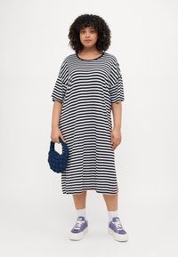 Randig, svart och vit oversized klänning med korta ärmar, sidoklipp, i kombination med en texturerad marinblå handväska och lila sneakers.