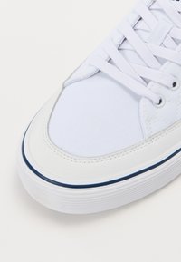 Detailný záber prednej časti bieleho teniska s bielymi šnúrkami, plátenným materiálom, gumovou podrážkou a tenkým námorníckym modrým pruhom.