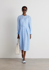 Light blue button-up dress with long sleeves, belted waist, and white polka dots. Paired with light blue slides and a beige handbag.