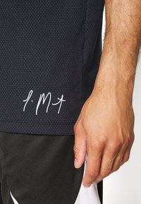 Námořnické sportovní tričko s texturovaným vzorem, zdobené bílými švy a logem v ručně psaném stylu na lemu, spárované s černými šortkami.