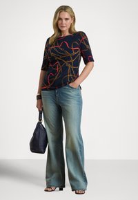 Žena v námořnicky modrém topu s potiskem červených a hnědých řetězů, světle modrých širokých džínách, černých sandálech, držící námořnicky modrou kabelku, pózující na bílém pozadí.