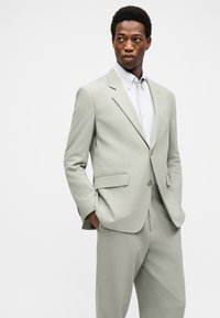 Άνδρας με ανοιχτό γκρι κοστούμι και λευκό πουκάμισο με κουμπιά, τα χέρια στις τσέπες, κοιτάζοντας ελαφρώς στο πλάι μπροστά σε απλό φόντο.