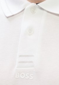 Λευκό πόλο μπλουζάκι με υφή υφάσματος, διαθέτει κούμπωμα με τρία κουμπιά, μικρό λογότυπο στο κάτω μπροστινό μέρος και διακριτικές οριζόντιες ρίγες.