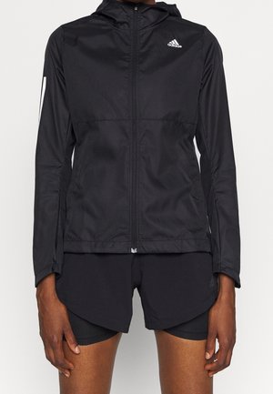 Close-up van een persoon in een zwarte Adidas zip-up jack en bijpassende zwarte sportshorts, staand tegen een effen achtergrond.