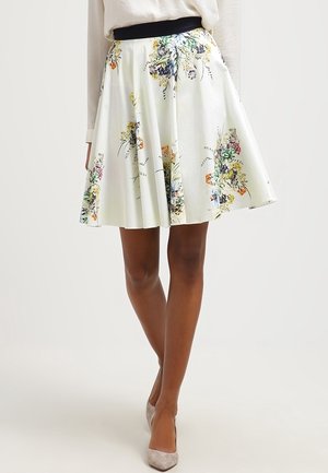 Femme portant une jupe blanche en ligne A avec des imprimés floraux colorés et une ceinture noire, associée à des talons pointus de couleur claire.