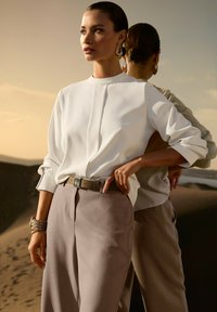 Frau in weißer Bluse und beigen Hosen steht mit einer Hand auf der Hüfte, großen Creolen und einem Armband vor einem sandigen Wüstenhintergrund bei Sonnenuntergang.