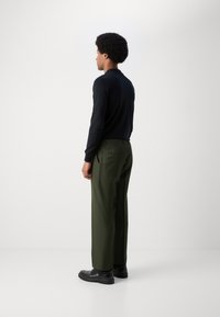 Chemise noire à manches longues, pantalon plissé vert olive, et chaussures noires. La tenue présente un design ajusté et une texture lisse.